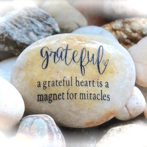 May include: A smooth, light brown stone with the words "grateful" and "a grateful heart is a magnet for miracles" engraved in black.