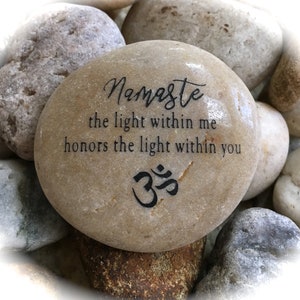 Op de afbeelding: Een gladde, lichtbruine steen met de woorden "Namaste the light within me honors the light within you" en het Om-symbool gegraveerd in zwart.