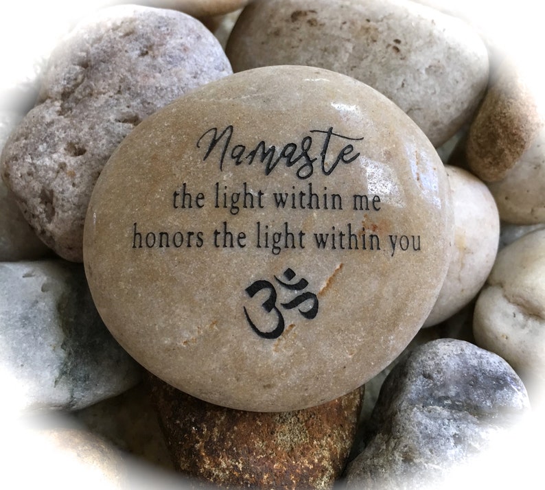 Op de afbeelding: Een gladde, lichtbruine steen met de woorden "Namaste the light within me honors the light within you" en het Om-symbool gegraveerd in zwart.