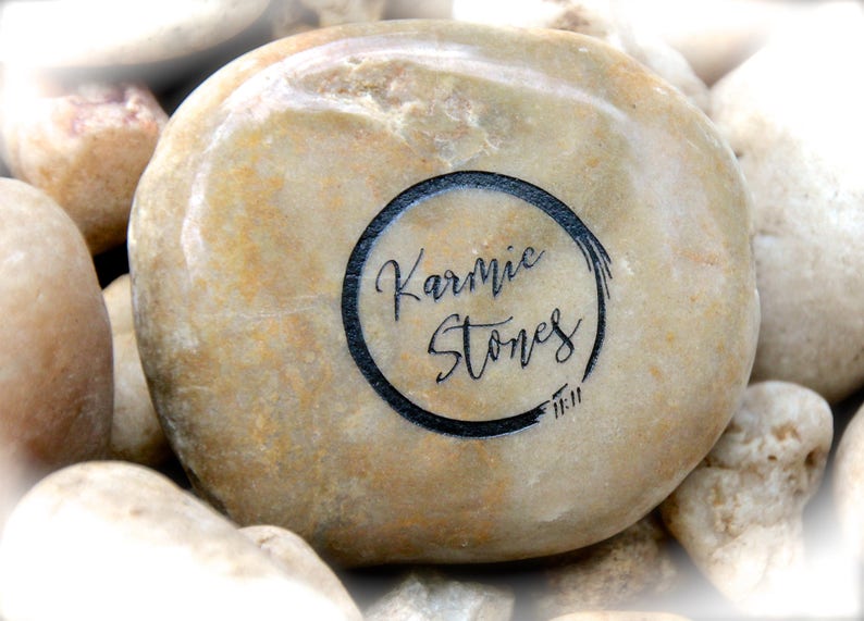 Op de afbeelding: Een close-up van een lichtbruine, gladde, ronde steen met een zwarte cirkel getekend rond de woorden "Karmic Stones" en de nummers "11:11" eronder geschreven.