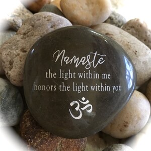 Op de afbeelding: Een donkergrijze steen met het woord "Namaste" en de zin "het licht in mij eert het licht in jou" erop gegraveerd. De steen heeft ook het Om-symbool gegraveerd.
