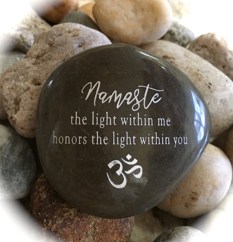 Op de afbeelding: Een donkergrijze steen met het woord "Namaste" en de zin "het licht in mij eert het licht in jou" erop gegraveerd. De steen heeft ook het Om-symbool gegraveerd.
