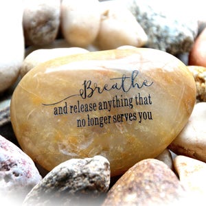 May include: A smooth, light brown stone with the words "Breathe and release anything that no longer serves you" inscribed in black.