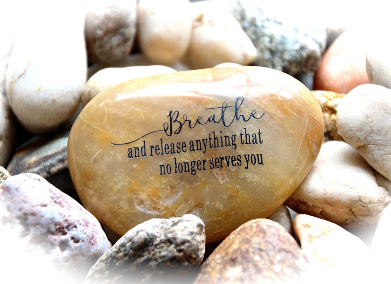 May include: A smooth, light brown stone with the words "Breathe and release anything that no longer serves you" inscribed in black.