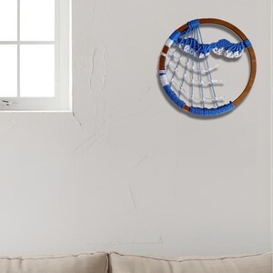 以下が含まれることがあります： 青と白の織り模様が施された、丸い木製のドリームキャッチャー。青と白のウェブデザインと雲のような装飾が特徴です。白い壁に掛けられています。