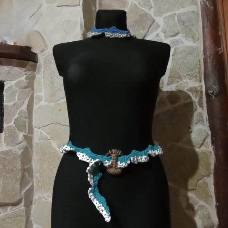 以下が含まれることがあります： 手作りのかぎ針編みセット。青と白のベルトにはロブスタークラスプ、お揃いのチョーカーが付いています。黒いマネキンに石壁を背景に展示されています。ベルトの長さは約76cmです。