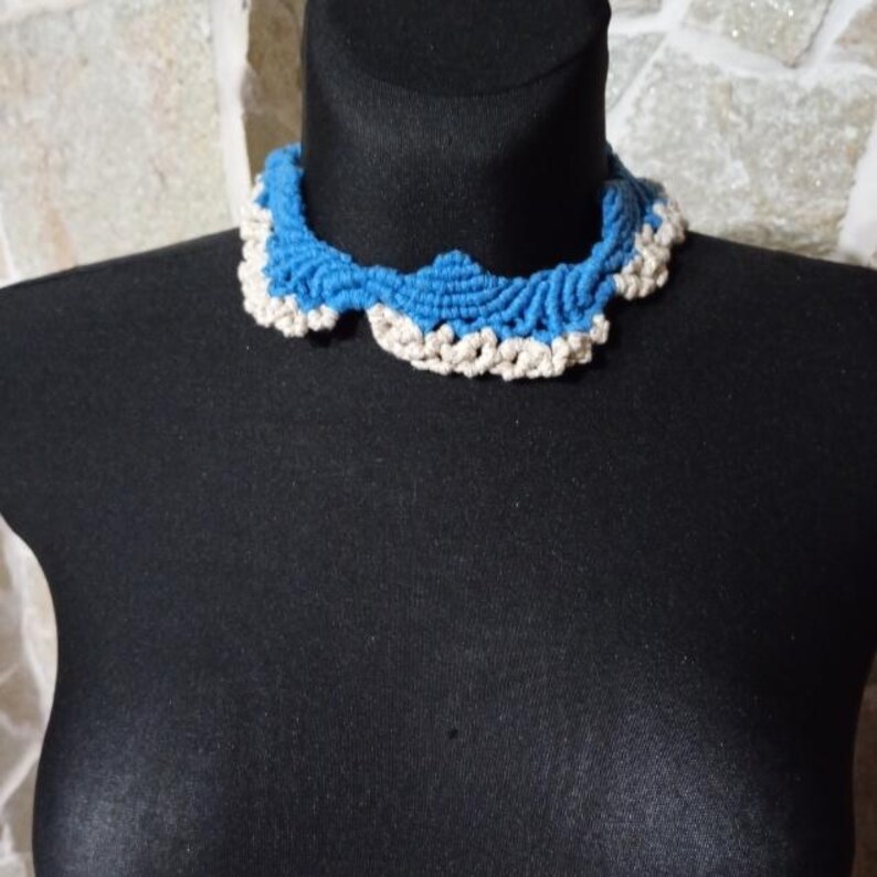 以下が含まれることがあります： 黒いマネキンに飾られた、青とクリーム色のクロシェカラーネックレス。 ネックレスはスカラップエッジとテクスチャードデザインが特徴で、手作りの美学を示しています。