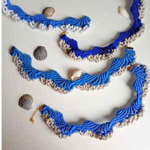 以下が含まれることがあります： 手作りの青とベージュのマクラメネックレス。波のようなデザインで、小さな貝殻が飾られています。複雑なマクラメの技術が、海をイメージしたテクスチャのある美しさを生み出しています。ゴールドカラーの留め金が付いています。