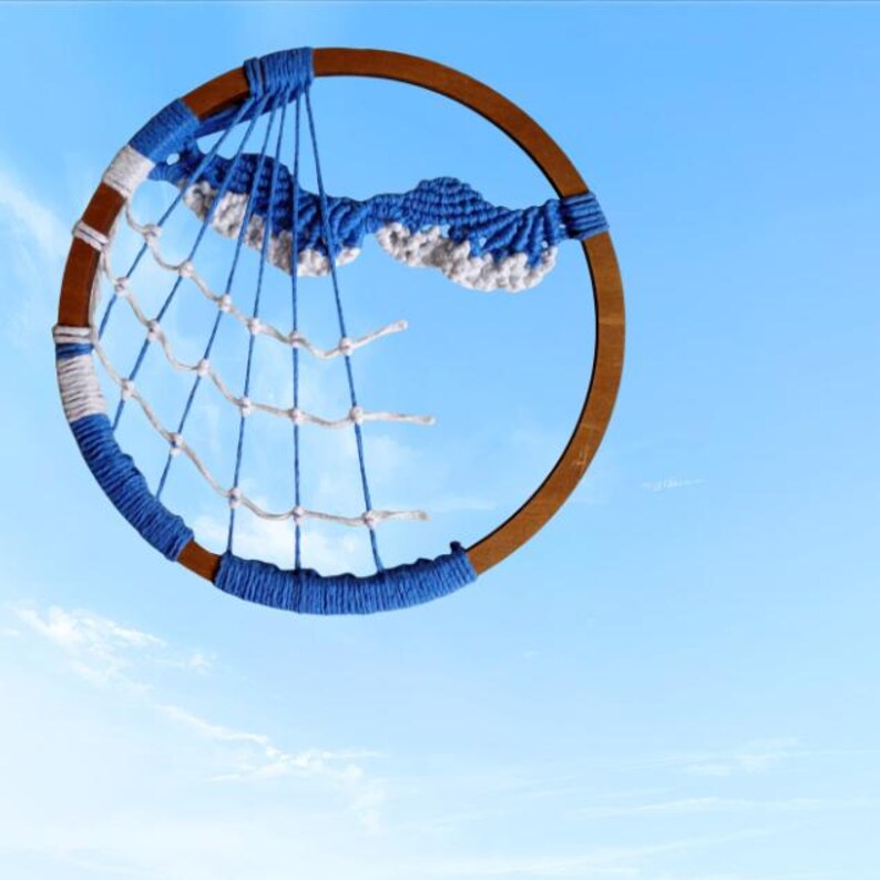 以下が含まれることがあります： 木製のフープと青と白の織りウェブデザインが特徴の手作りのドリームキャッチャー。 ドリームキャッチャーは、青と白の糸のアクセントと雲のような装飾が特徴で、青い空を背景にしています。