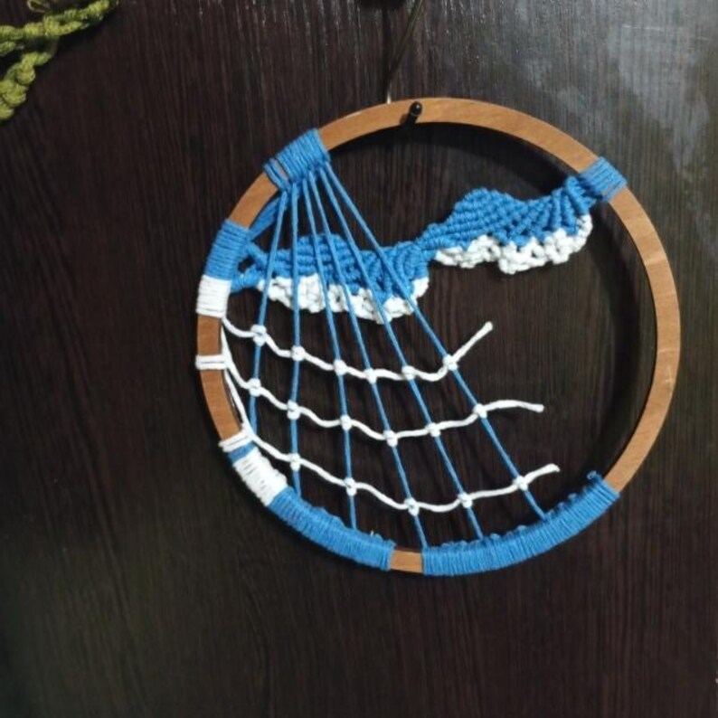 以下が含まれることがあります： 木製のフープが付いた手作りのドリームキャッチャー。フープは青い糸で包まれ、内側には白と青のウェブデザインがあります。上部には青と白の雲のようなデザインがあります。ドリームキャッチャーは濃い木材の背景に吊るされています。