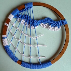 以下が含まれることがあります： 木製の輪で作られた手作りのドリームキャッチャー。白い糸と青い縦糸で織られたウェブパターンが特徴です。上部には青と白の雲の形をしたデザインがあります。輪は青い糸で包まれています。
