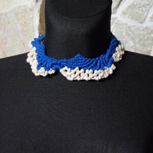 以下が含まれることがあります： 手作りの青とオフホワイトのかぎ針編みの襟ネックレス。ネックレスは黒いマネキンに展示されています。襟はスカラップエッジとテクスチャードデザインが特徴で、複雑な職人技を示しています。色の組み合わせは印象的なコントラストを生み出しています。