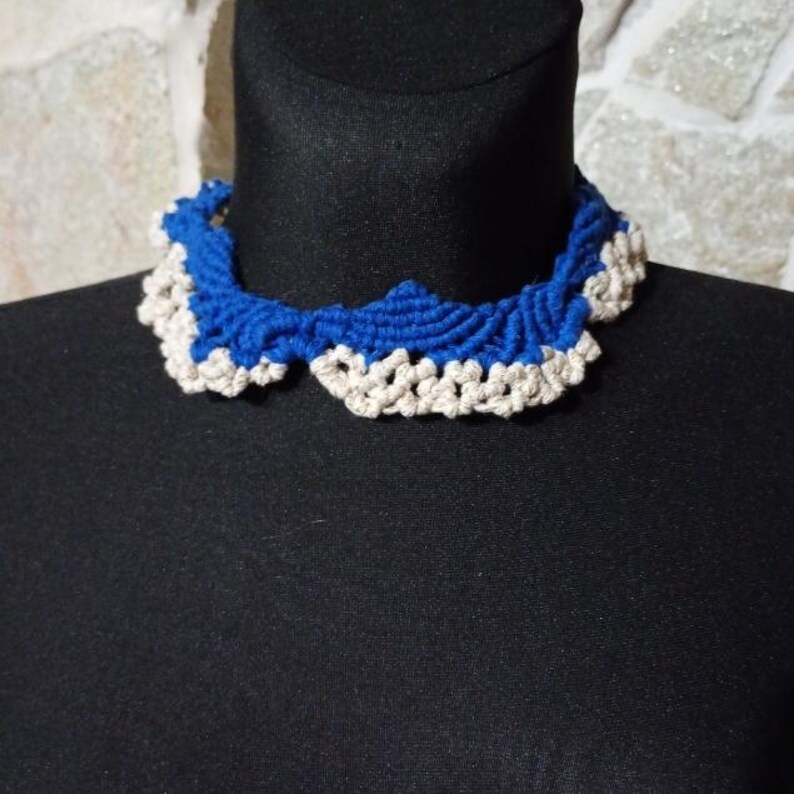 以下が含まれることがあります： 手作りの青とオフホワイトのかぎ針編みの襟ネックレス。ネックレスは黒いマネキンに展示されています。襟はスカラップエッジとテクスチャードデザインが特徴で、複雑な職人技を示しています。色の組み合わせは印象的なコントラストを生み出しています。
