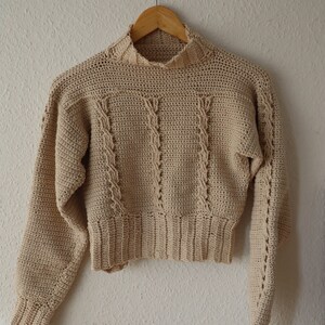 Puede incluir: Un suéter de punto de ganchillo beige con cuello alto y mangas largas. El suéter tiene un patrón de punto de cable y una longitud recortada.