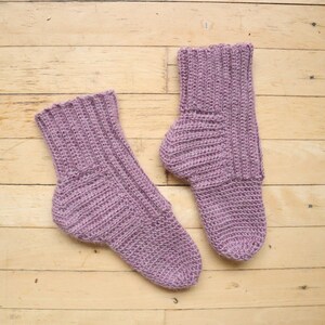 May include: Two pairs of mauve knitted socks with a ribbed texture. The socks are laid flat on a wooden surface.