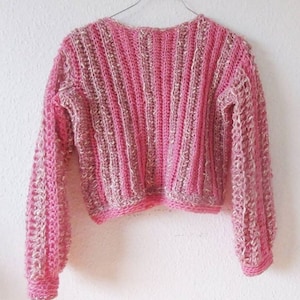 May include: A pink and beige crochet sweater with long sleeves. The sweater has a ribbed design on the body and sleeves, with a slightly cropped fit. The sleeves have a textured, openwork pattern.
