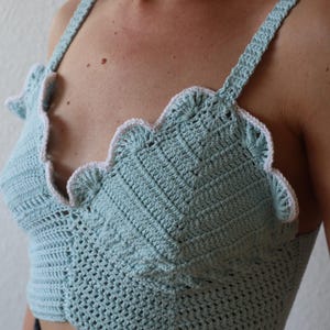 May include: A light blue crochet top with a scalloped edge and straps. The top is handmade with a detailed stitch pattern and a white trim along the edges. The top is a crop top style.