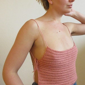 May include: A pink crocheted tank top with thin straps and a loose fit. The top has a square neckline and is tied at the sides.
