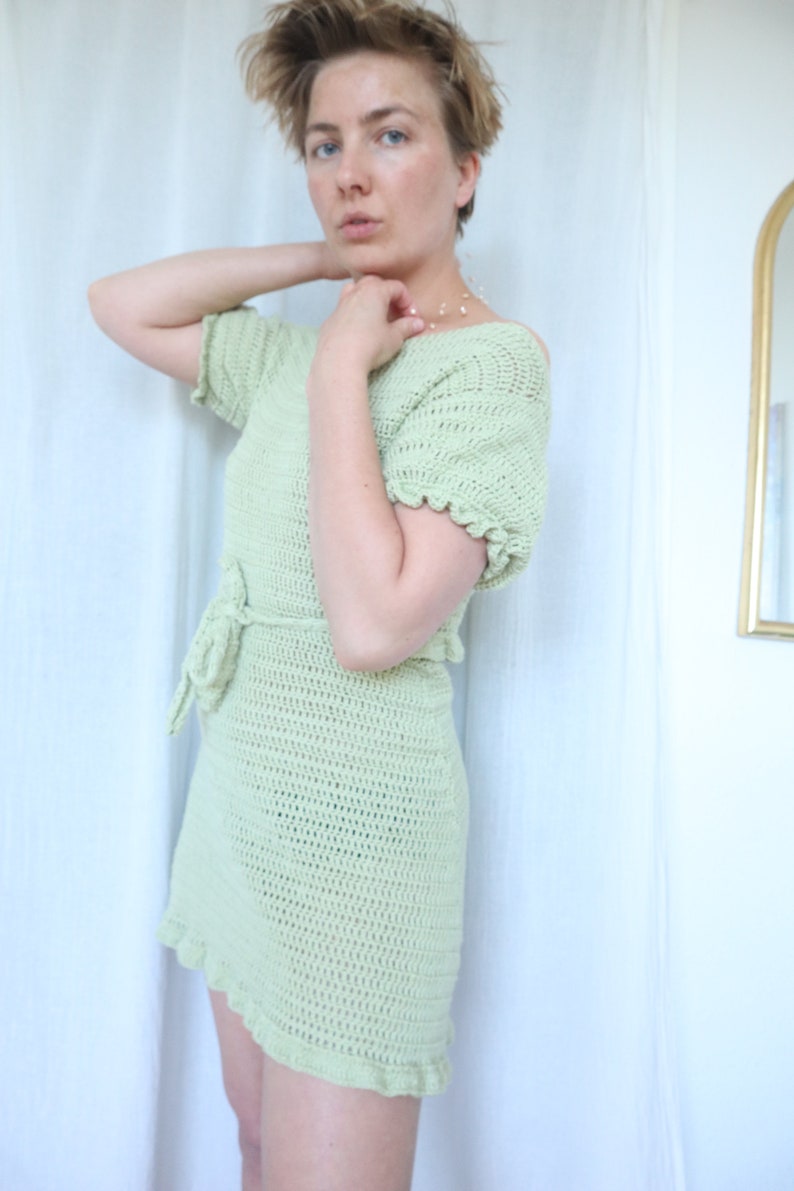May include: A light green crochet dress with a tie belt and short sleeves. The dress has a scalloped hem and a relaxed fit.