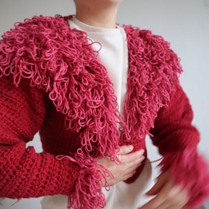 May include: A cropped pink cardigan with a fuzzy, crocheted texture. The cardigan has a V-neck and long sleeves with fringe detailing.