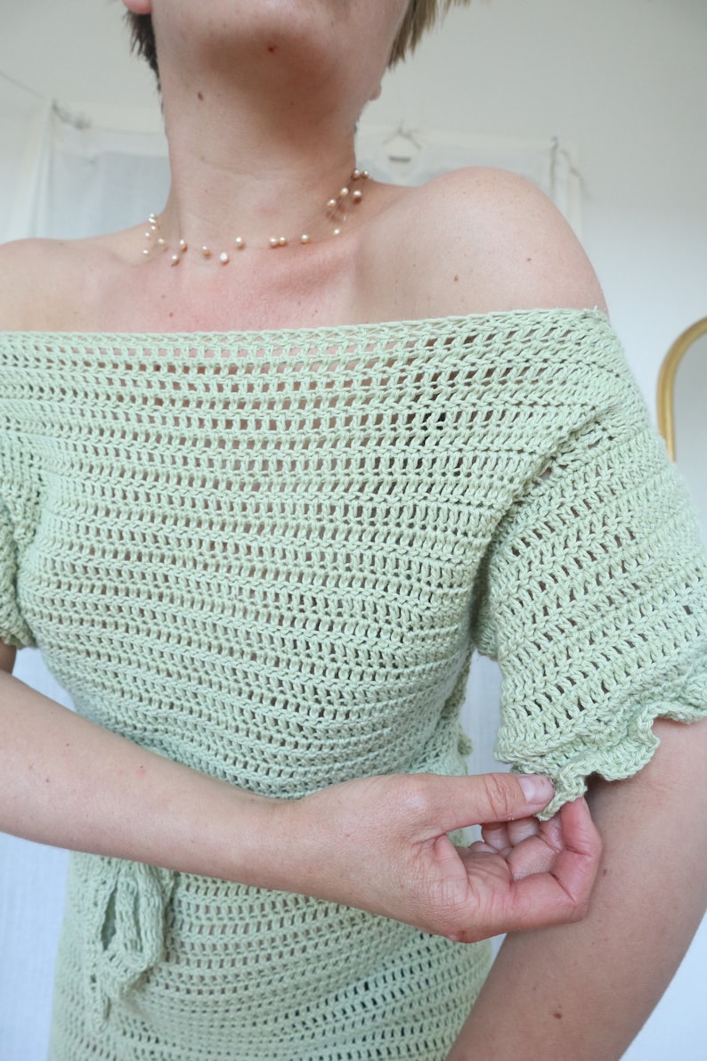 May include: A light green crochet top with a loose, off-the-shoulder fit and short sleeves. The top has a delicate, open-weave pattern and a tie at the waist.