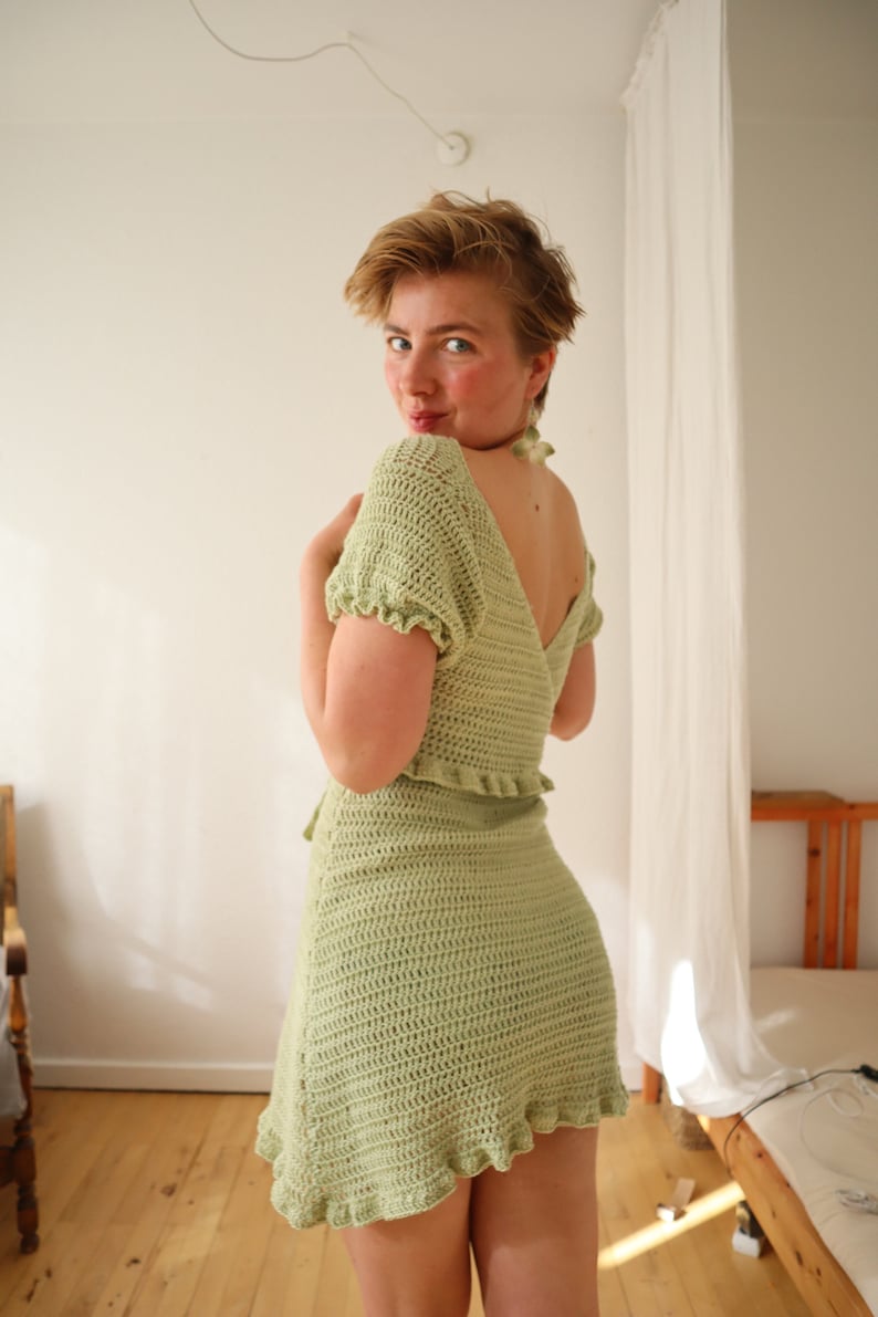 May include: A light green crochet dress with a ruffled hem and short sleeves. The dress is fitted and has a V-neckline.