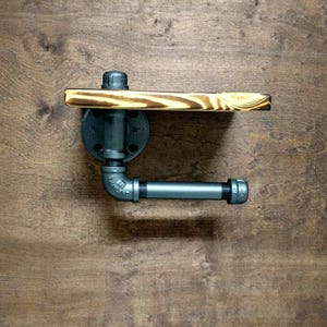 May include: Industrial toilet roll holder made from black metal pipes and a wooden shelf. The wood has a natural grain pattern. The holder is mounted on a wooden surface.