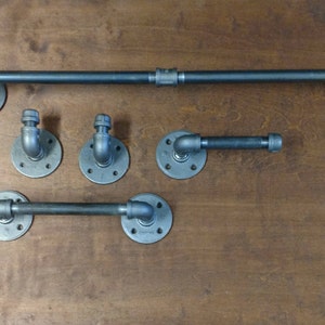 May include: A set of industrial-style bathroom fixtures, including a towel bar, two towel hooks, and a toilet paper holder. The fixtures are made of dark gray metal pipes and fittings, mounted on circular flanges.