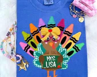 May include: A blue t-shirt featuring a graphic of a brown turkey with a yellow bow and pink glasses. The turkey's tail feathers are made of colorful crayons. A green chalkboard sign held by the turkey reads Mrs. LISA. A gold star necklace and a pink hair clip are included.