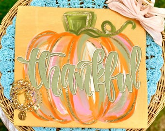 May include: A mustard yellow t-shirt featuring a large, painterly orange pumpkin graphic with a green stem and the word thankful written in cursive script across the center. A stack of gold-toned beaded bracelets with a cross charm sits on the shirt.