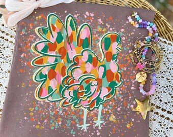May include: A brown fabric square featuring a turkey graphic filled with multicolored brushstrokes in shades of orange, green, and pink. The turkey is surrounded by paint splatters and a light pink ribbon bow is attached to the top corner.