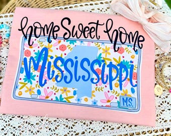 Puede incluir: Una camiseta rosa claro con un gráfico de una matrícula con estampado floral, el texto "home sweet home" en negro y la palabra "Mississippi" en letra azul. Se observa un contorno de estado en azul claro y la abreviatura "MS.".