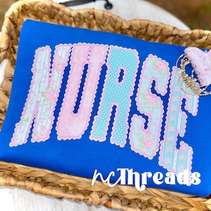 May include: A royal blue shirt with the word "NURSE" in large, decorative letters. The letters are in pink, light blue, and floral patterns. The text "nCThreads" is at the bottom. The shirt is displayed in a woven basket.
