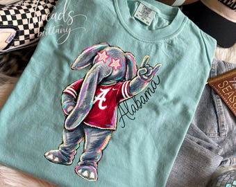 May include: A light mint green t-shirt featuring a graphic of a gray elephant wearing a crimson jersey with a white letter A. The elephant has pink star-shaped eyes and is making a hand gesture. The word Alabama is written in black script.
