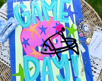Puede incluir: Una camiseta azul con el texto GAME DAY en letras grandes de color turquesa. El diseño incluye un gráfico circular rosa con estrellas naranjas, azules y verdes, y el contorno de un casco de fútbol americano negro. Una pinza de pelo a cuadros y pulseras.