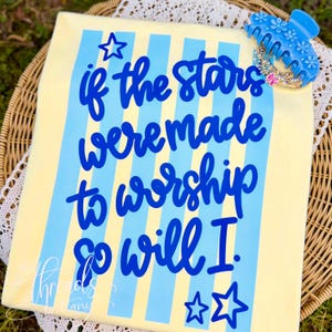 May include: A light yellow t-shirt with blue vertical stripes and the text "if the stars were made to worship so will I" in blue script. A blue hair clip and a small gold crown are on the top right.