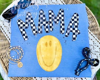Puede incluir: Una camiseta azul con la palabra MAMA en un patrón de cuadros blancos y negros con salpicaduras de pintura dorada. Debajo del texto hay una cara sonriente amarilla grande. La camiseta se muestra con gafas de sol negras, una pulsera dorada y una pinza para el pelo negra.
