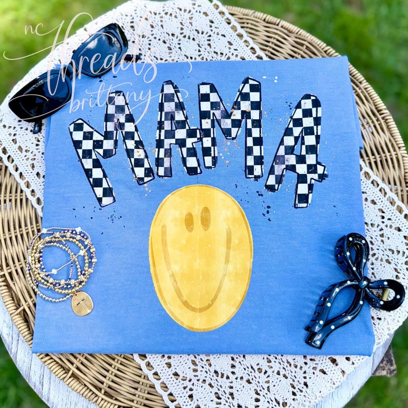 K&ouml;nnte beinhalten: Ein blaues T-Shirt mit dem Wort MAMA in einem schwarz-wei&szlig;en Karomuster und goldenen Farbspritzern. Unter dem Text befindet sich ein gro&szlig;es gelbes Smiley-Gesicht. Das T-Shirt wird mit einer schwarzen Sonnenbrille, einem goldenen Armband und einer schwarzen Haarspange pr&auml;sentiert.