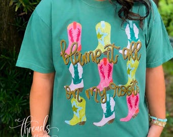 Puede incluir: Una camiseta de manga corta color verde azulado con un estampado gráfico de nueve botas vaqueras coloridas en rosa, amarillo y blanco. El texto "blame it all on my roots" está impreso en una fuente dorada estilo cuerda en el centro.