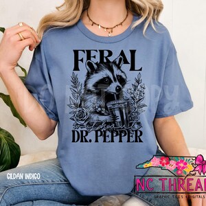 Puede incluir: Una camiseta azul con el texto "Feral" y "Dr. Pepper" impreso en ella. Una ilustraci&oacute;n en blanco y negro de un mapache bebiendo de una taza est&aacute; en el centro de la camiseta.