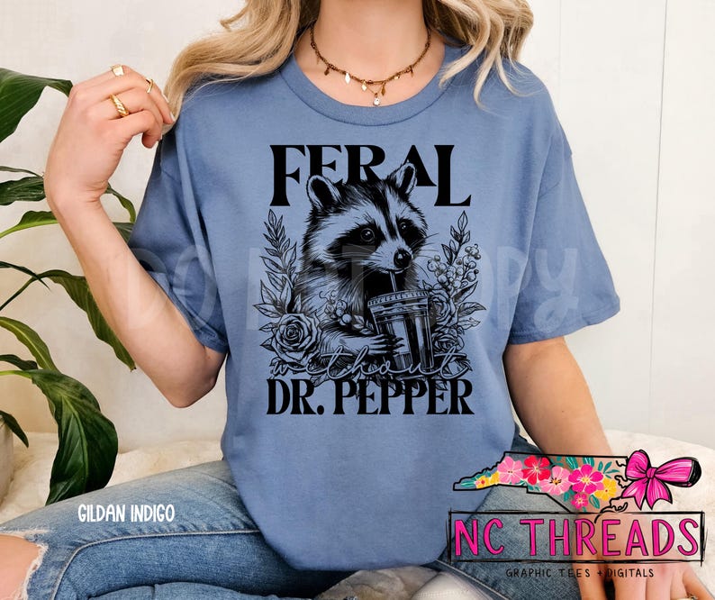 Puede incluir: Una camiseta azul con el texto "Feral" y "Dr. Pepper" impreso en ella. Una ilustraci&oacute;n en blanco y negro de un mapache bebiendo de una taza est&aacute; en el centro de la camiseta.