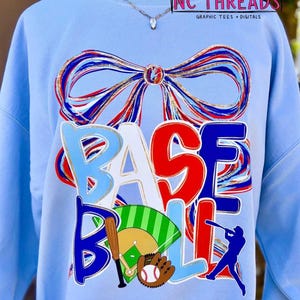May include: Light blue sweatshirt with a baseball theme. The word "BASEBALL" is displayed in a colorful, layered design. A bow graphic is at the top. Includes a baseball, bat, glove, and batter silhouette.