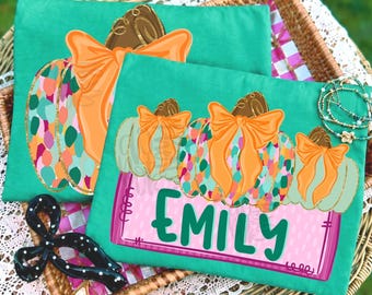 May include: Two teal t-shirts featuring a graphic of three pumpkins with orange bows. The pumpkins have a colorful speckled pattern. Below the pumpkins, a pink rectangular box contains the name EMILY in large, dark green capital letters.