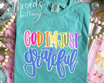 Puede incluir: Una camiseta de manga corta color verde azulado con el texto "GOD I'M JUST grateful" en un estilo manuscrito multicolor. La palabra "GOD" es rosa, "I'M" es naranja, "JUST" es amarillo y verde, y "grateful" está en una letra azul grande.