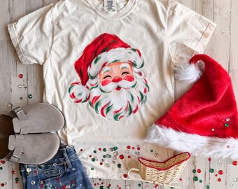 May include: A cream-colored short-sleeve t-shirt featuring a painted illustration of Santa Claus with a red hat and white beard. The shirt is displayed with a red plush Santa hat, a small woven wicker sleigh, and grey suede clogs on blue jeans.