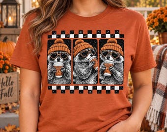 May include: A burnt orange short-sleeve t-shirt featuring a graphic print of three raccoons wearing orange beanies and heart-shaped sunglasses. Each raccoon holds a coffee cup labeled Pumpkin Spice, framed by a black and white checkered border.
