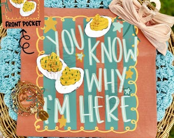 May include: A terracotta-colored shirt featuring a teal and red striped background with yellow stars and illustrations of deviled eggs. The text reads, "YOU KNOW WHY I'M HERE." A light pink bow is attached to the top right, and a gold bracelet is on the left.