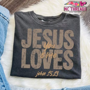 May include: Dark gray t-shirt with the text "JESUS loves you & me" in distressed brown lettering. The Bible verse John 15:13 is printed below the text. The shirt is folded on a white fluffy surface.