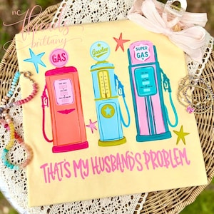 May include: A light yellow t-shirt featuring three vintage gas pumps in pink, blue, and teal, with star accents. The text "THAT'S MY HUSBAND'S PROBLEM" is written in pink script across the bottom. A fun, retro-themed graphic tee.