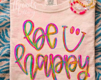 Puede incluir: Una camiseta de color beige claro con el texto "be happy" en una vibrante escritura de neón multicolor y un icono de cara sonriente. La camiseta tiene una etiqueta Comfort Colors en el cuello y se muestra con pulseras de cuentas.